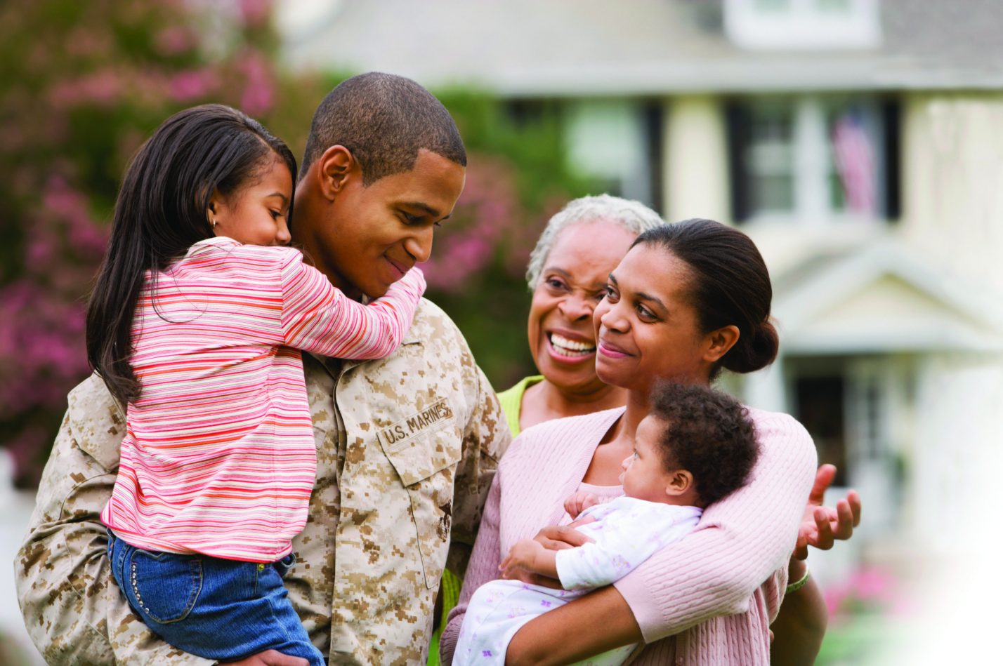 A multi-generational family stands huddled together in front of a two-story home. The father is wearing service fatigues.