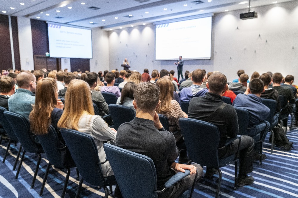 Man speaking on a stage to a large audience in a conference room