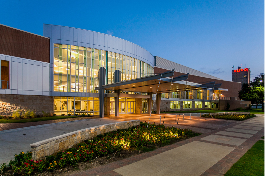 A large building is featured at dusck. In the background stands a larger building flying the American flag and having the title "Alico".