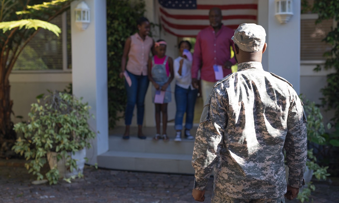 Father in uniform approaches his wife, children who are greeting him outside the house.